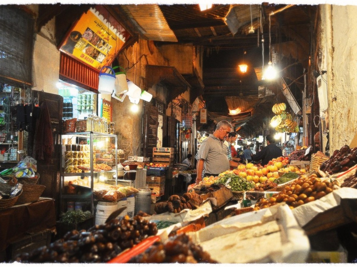 Indoor market scene with stalls selling fruits, vegetables, and dried goods, and people shopping.