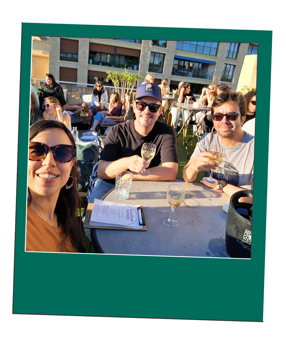 Three people enjoying drinks at an outdoor cafe with others in the background.