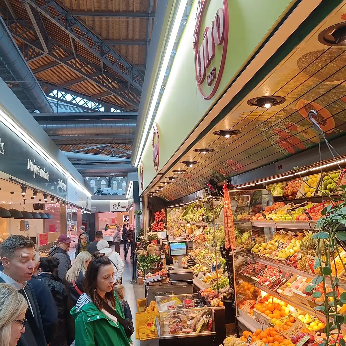 Indoor market with people shopping at a vibrant produce stall filled with fruits and vegetables.