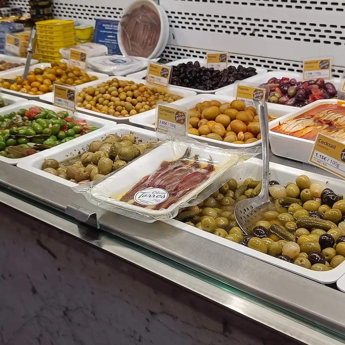 Display of assorted olives and pickles in a market with price labels.