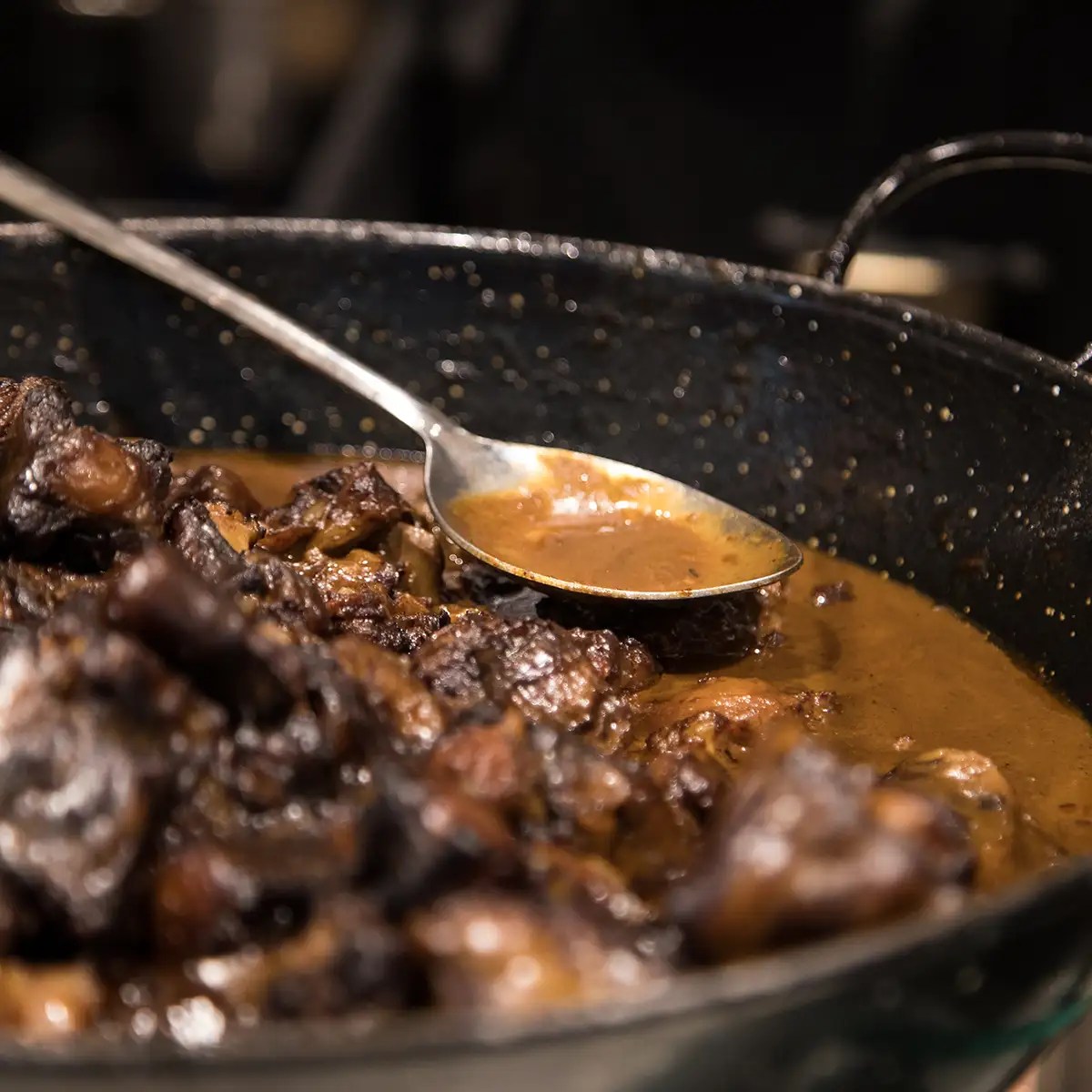 Close-up of a spoon in a pan with dark meat and brown sauce.
