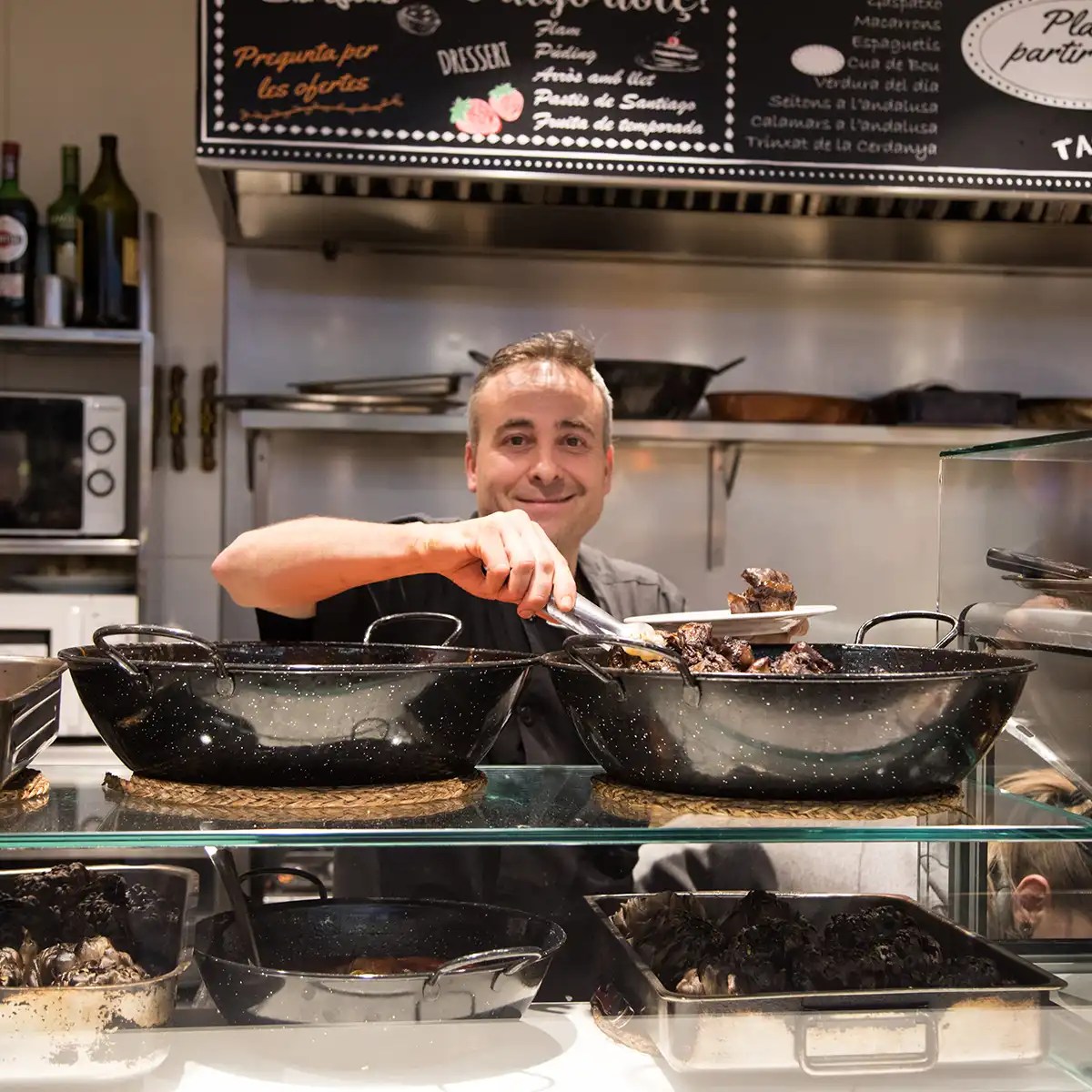 Chef serving food from large black pots in a commercial kitchen with a menu board above.