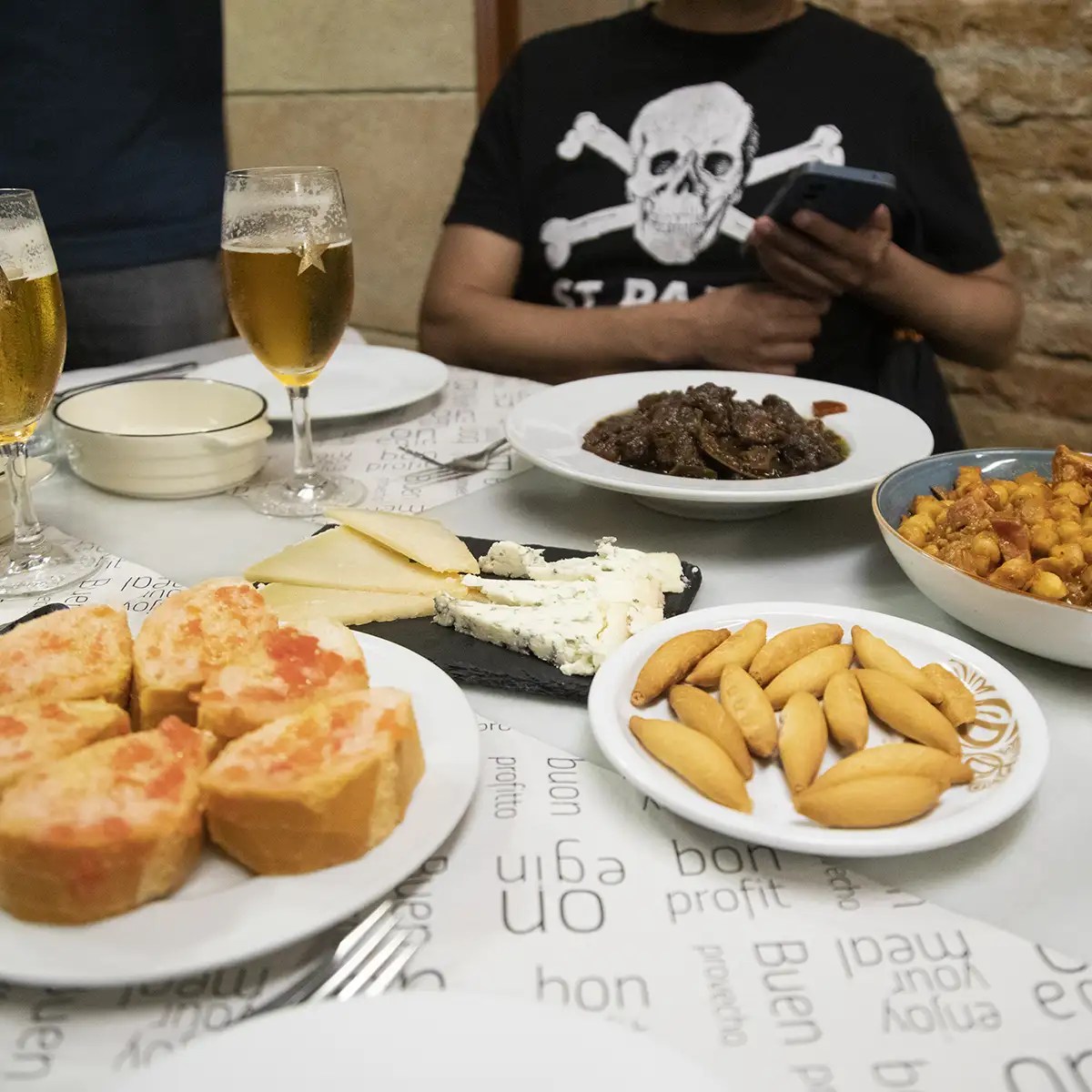 Table with various dishes, cheeses, bread, and beer glasses, person in skull t-shirt in background.
