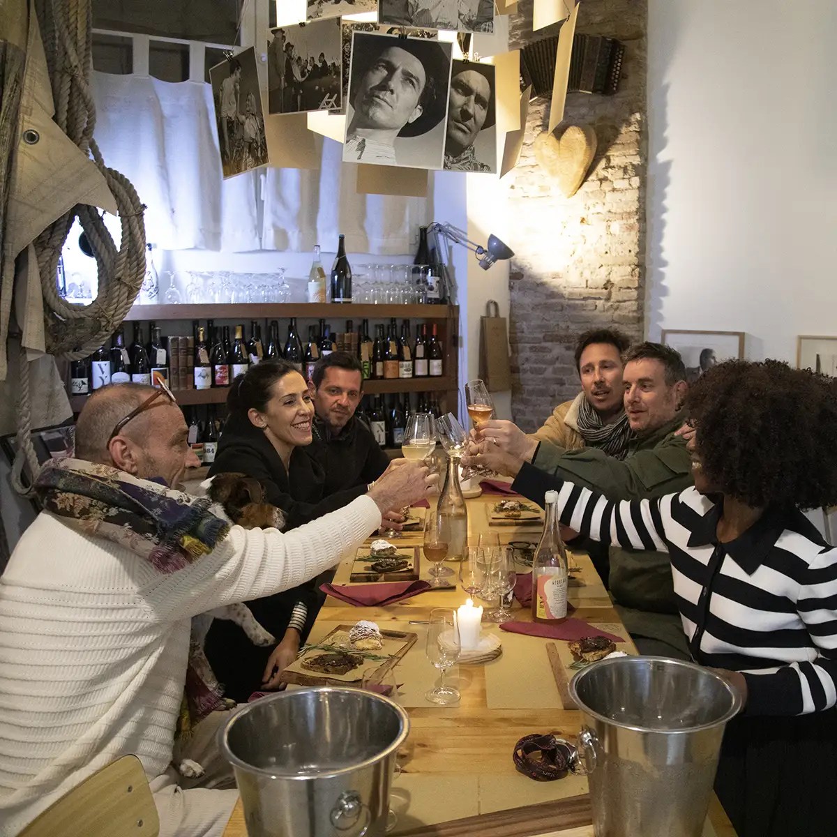Group of people toasting with drinks at a cozy dinner table with wine bottles and hanging photos.
