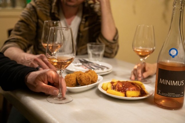 People dining with wine glasses, fried snacks, and a bottle labeled 'MINIMUS' on a table.