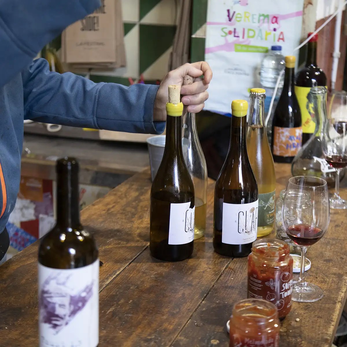 Person opening wine bottle at wooden table with jars and wine glasses nearby.