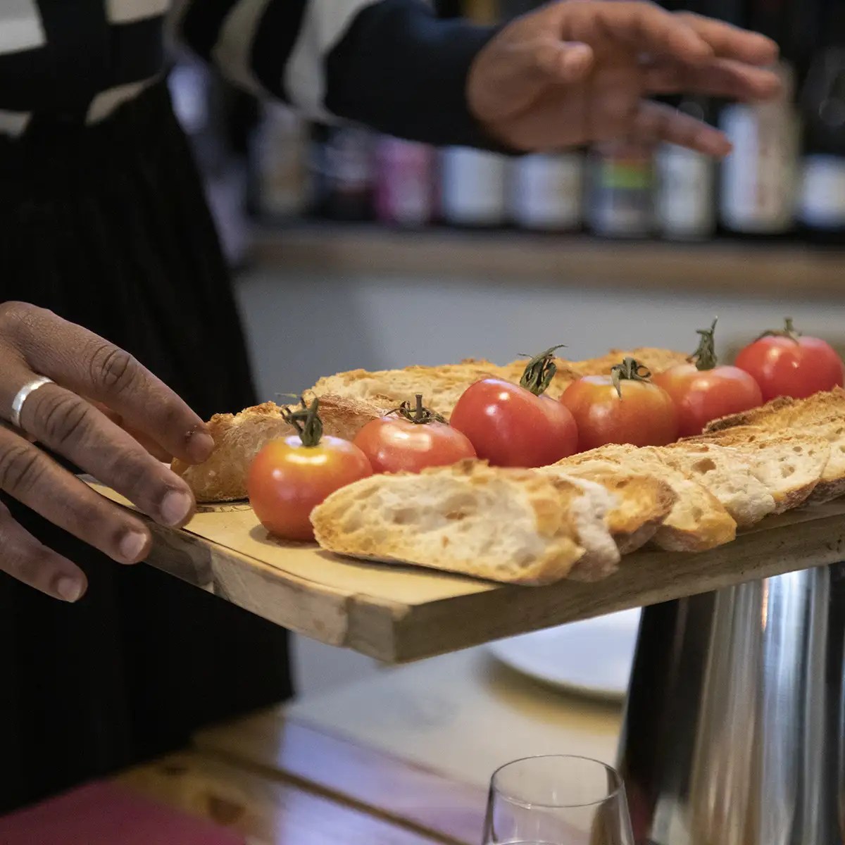 Hands plating bread and tomatoes on a wooden board in a kitchen setting.