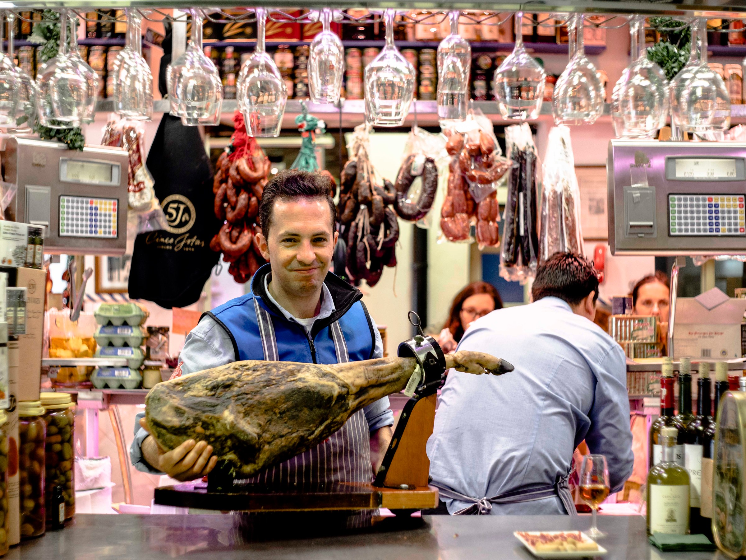 Man in a deli holding a leg of ham, with sausages and wine bottles displayed around him.