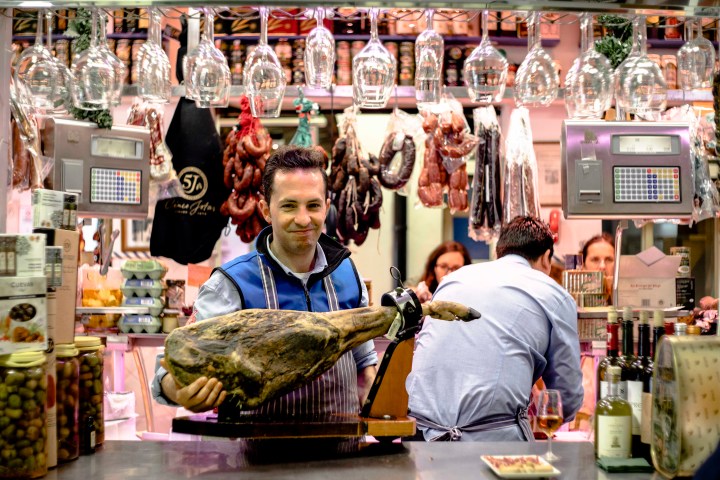 Man in a deli holding a leg of ham, with sausages and wine bottles displayed around him.