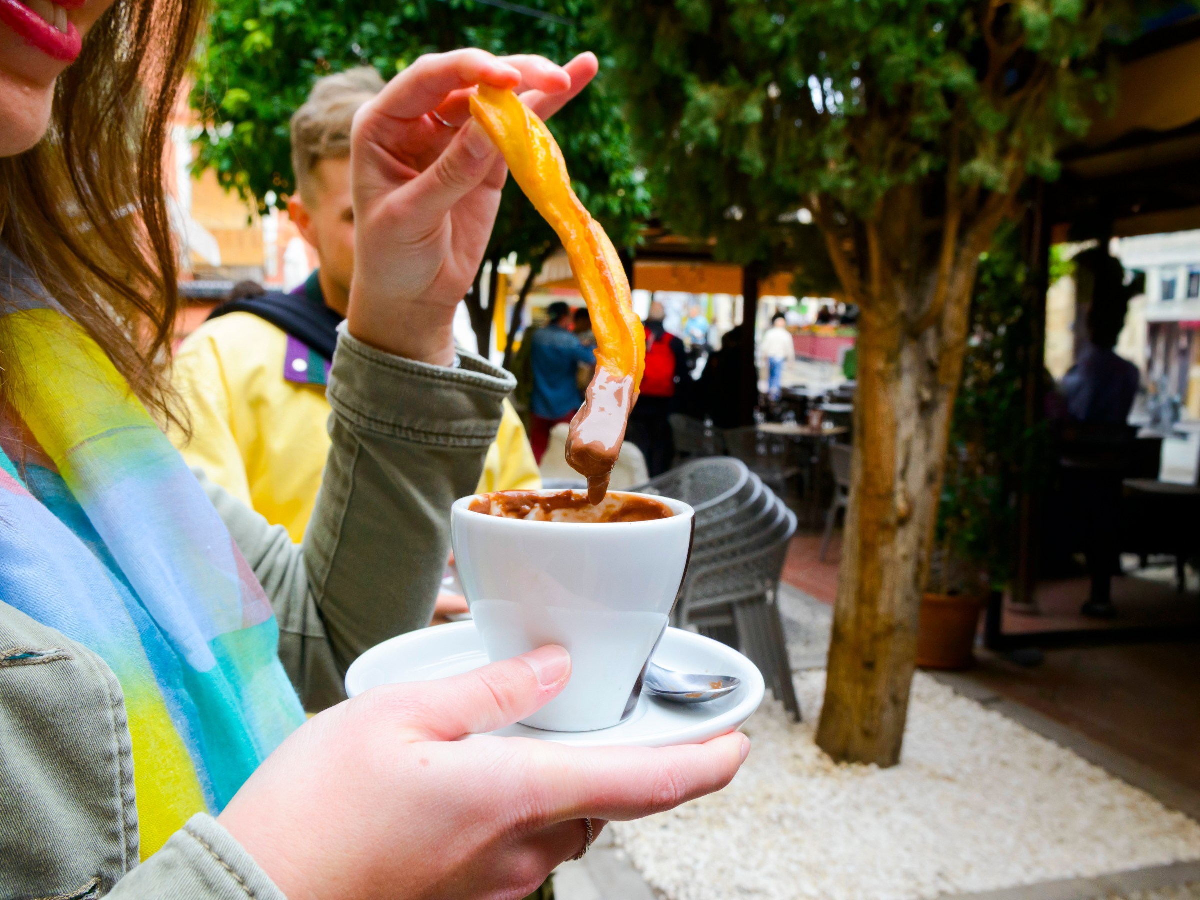 Person holding churro dipped in chocolate at an outdoor café.