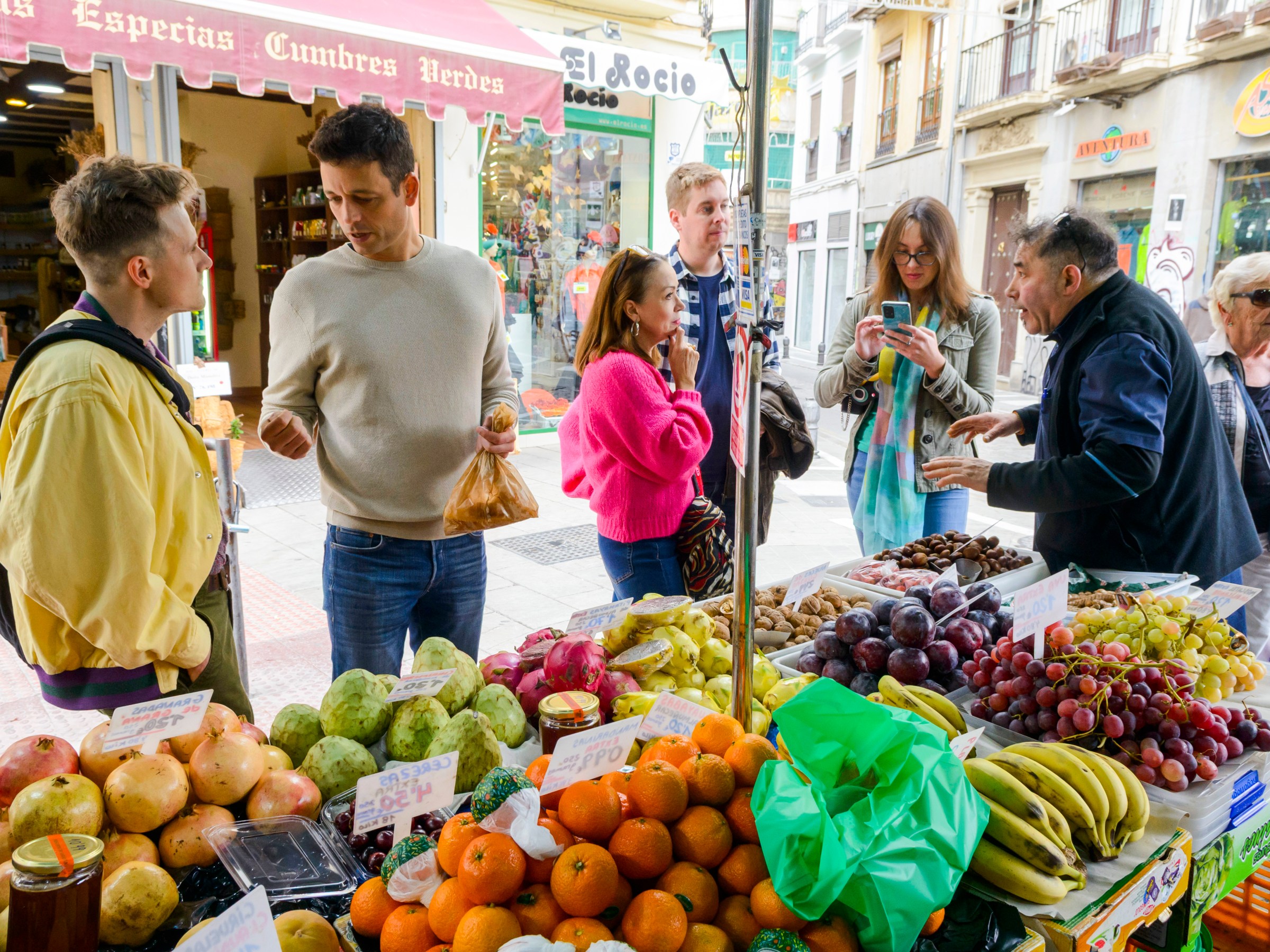 People shopping at an outdoor fruit stall with various fresh produce in a busy market.