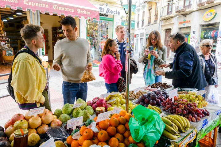People shopping at an outdoor fruit stall with various fresh produce in a busy market.