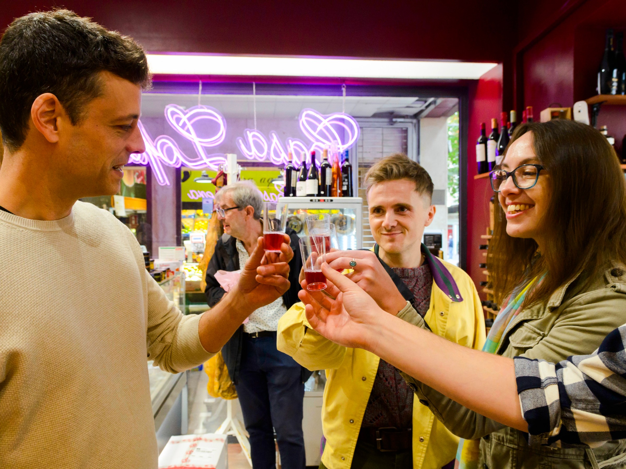 Three people clinking glasses in a store with a neon sign in the background.