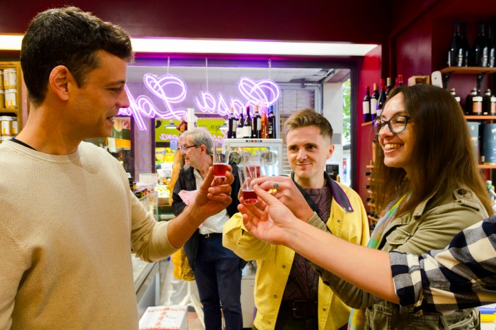 Three people clinking glasses in a store with a neon sign in the background.