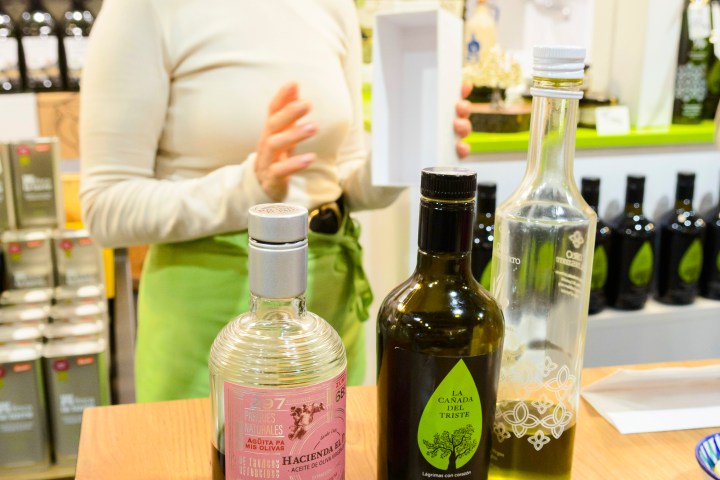 Woman presenting olive oils on a wooden counter with various bottles visible.
