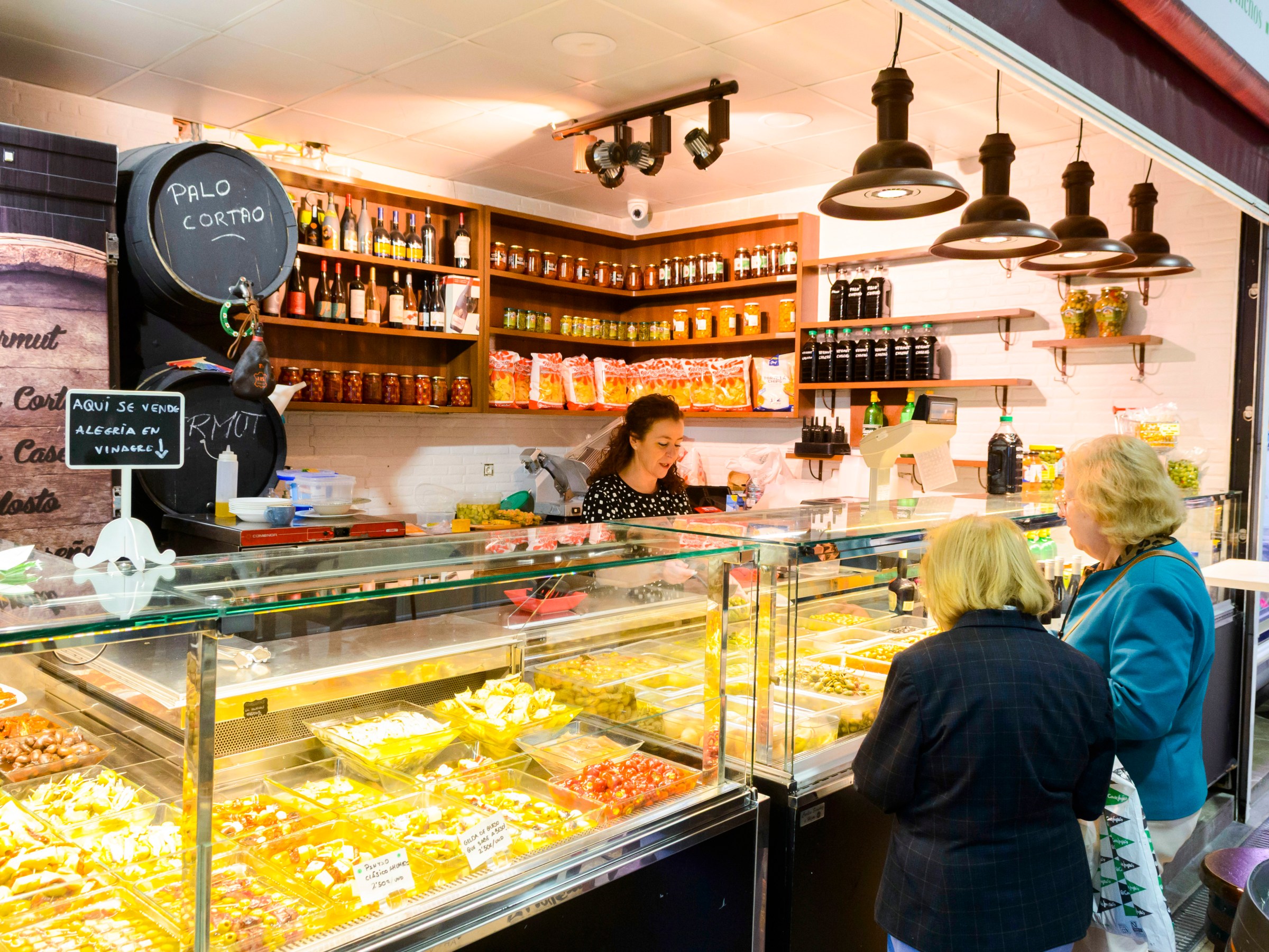 Market stall with various pickles and jars, served by a woman to two customers.