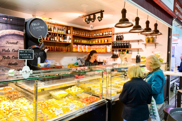 Market stall with various pickles and jars, served by a woman to two customers.