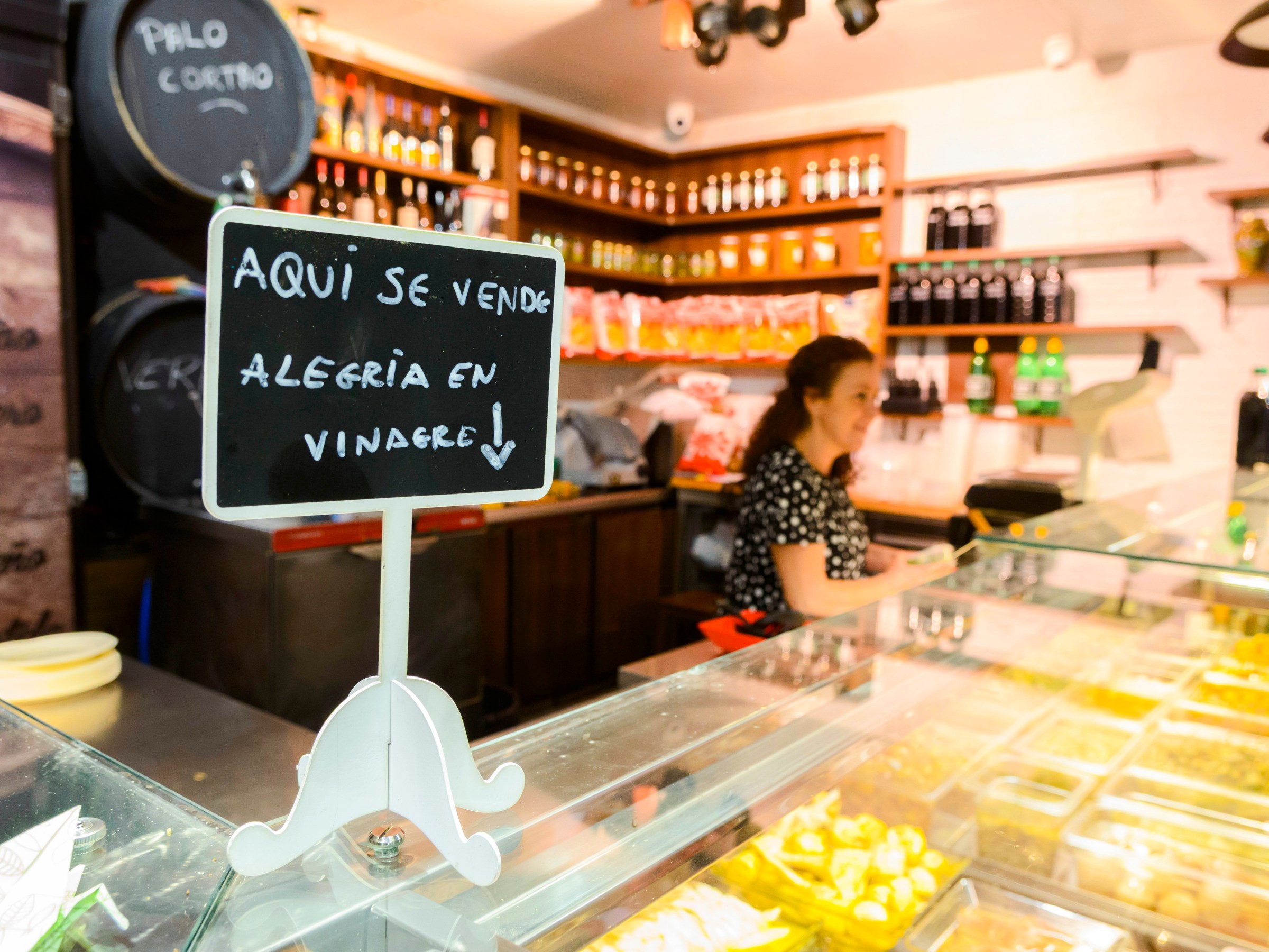 Woman behind deli counter with sign reading 'Aquí se vende alegría en vinagre' in Spanish.