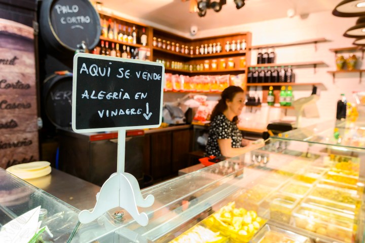 Woman behind deli counter with sign reading 'Aquí se vende alegría en vinagre' in Spanish.