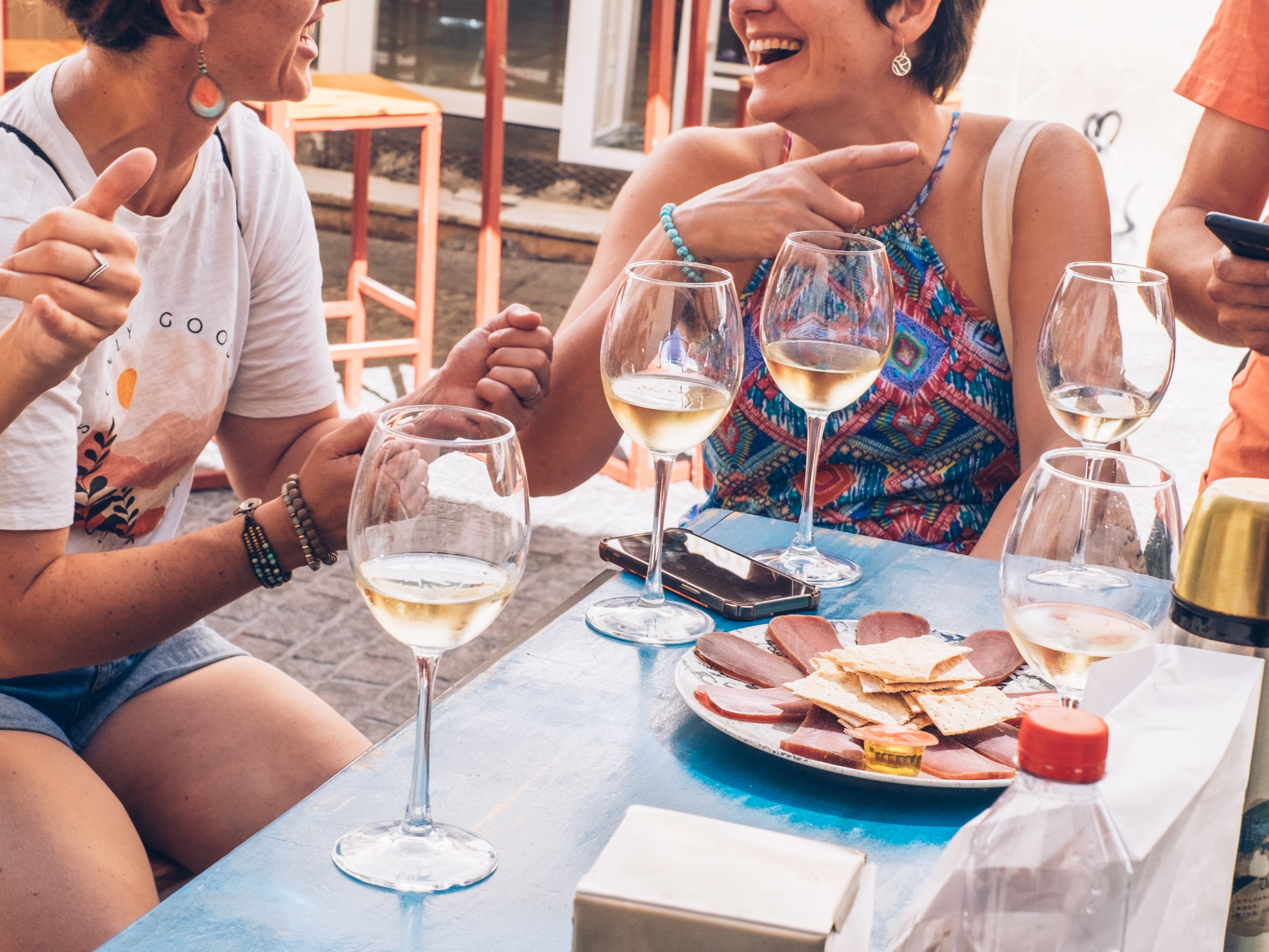 Two women laughing and enjoying wine at an outdoor table with snacks.