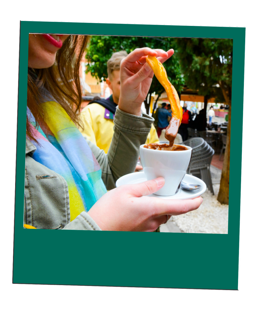 Person dipping pastry in chocolate cup outdoors, smiling with colorful scarf.