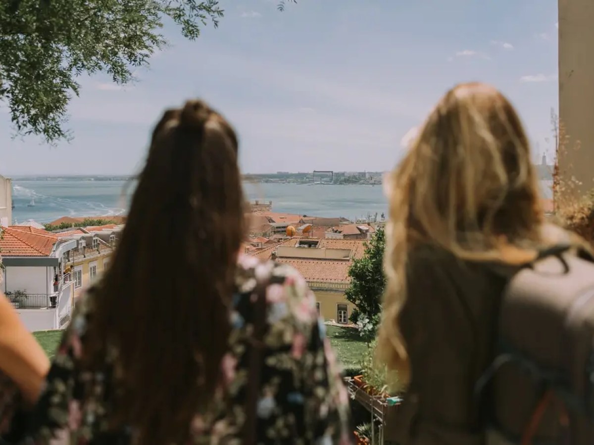 Two women with long hair overlook a scenic coastal city view.