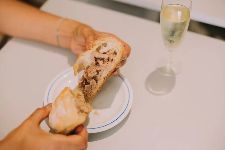 Person holding bread with meat filling over a plate beside a glass of champagne.